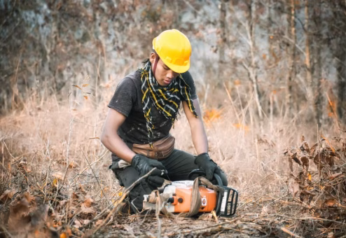Worker in helmet and protective chaps inspecting a chainsaw before cutting