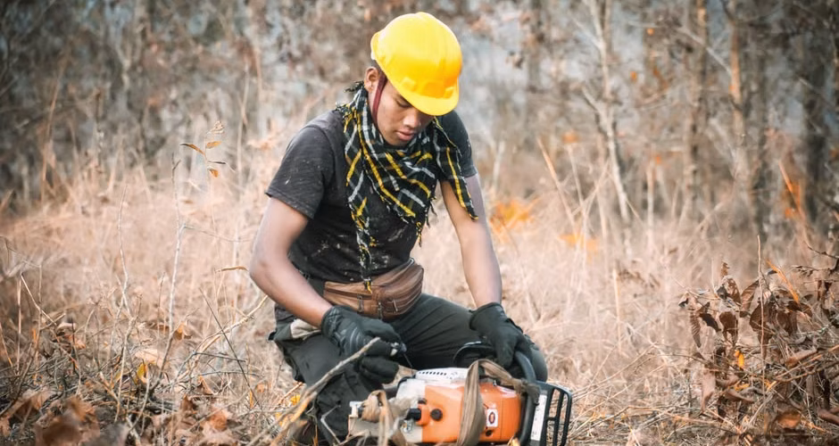 Worker in helmet and protective chaps inspecting a chainsaw before cutting